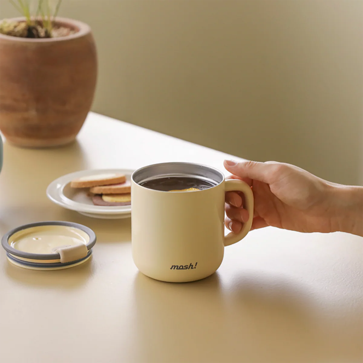 Hand holding a beige mug with 'mash!' branding on a table with a plant and cookies in the background.